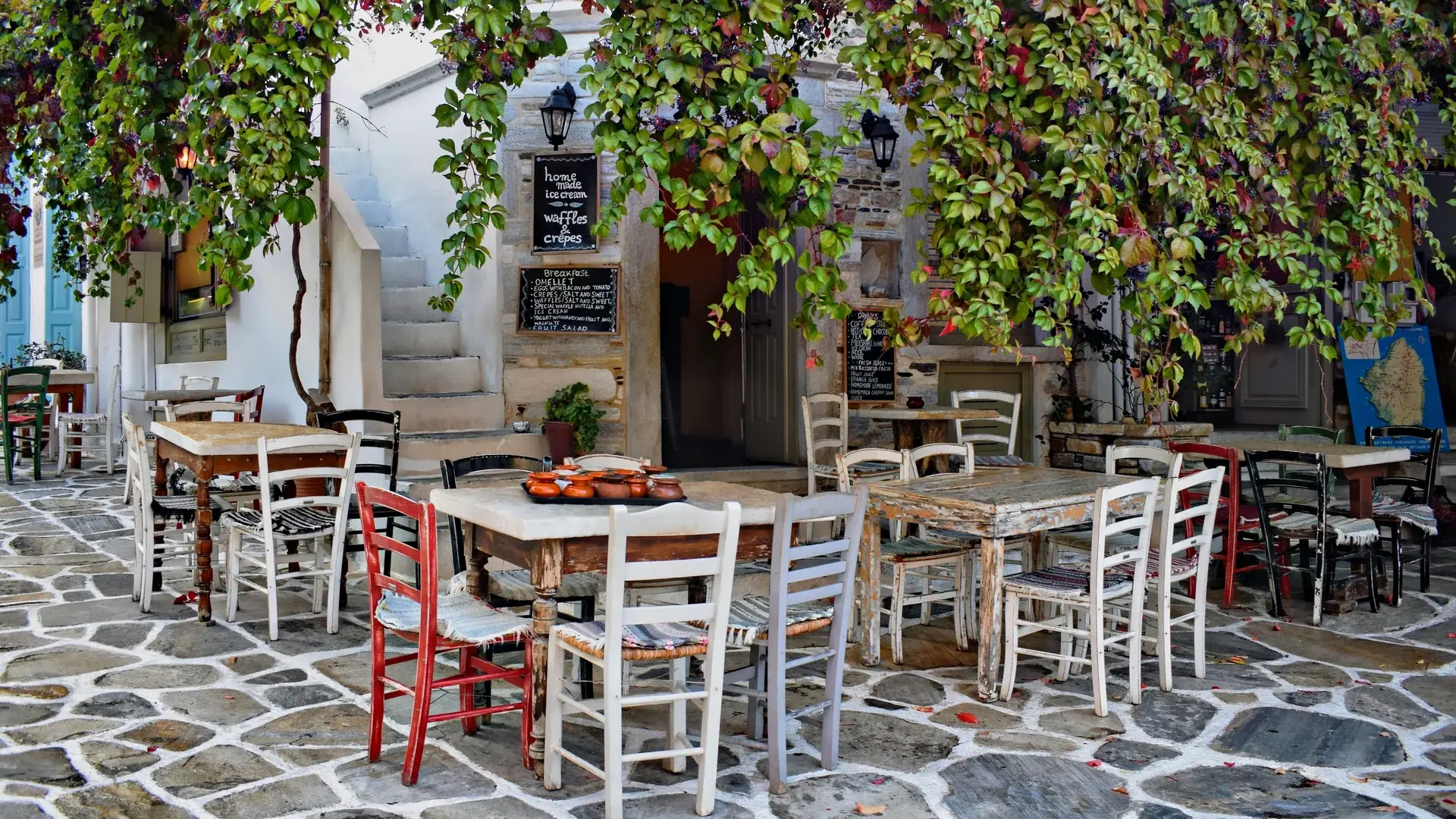 Vesnická taverna na Naxosu s nepasujícími dřevěnými stoly a červenobílými židlemi na dlážděném náměstí, ve stínu loubí z vinné révy, tác s hliněnými nádobami na jednom stole a mapa ostrova na zdi